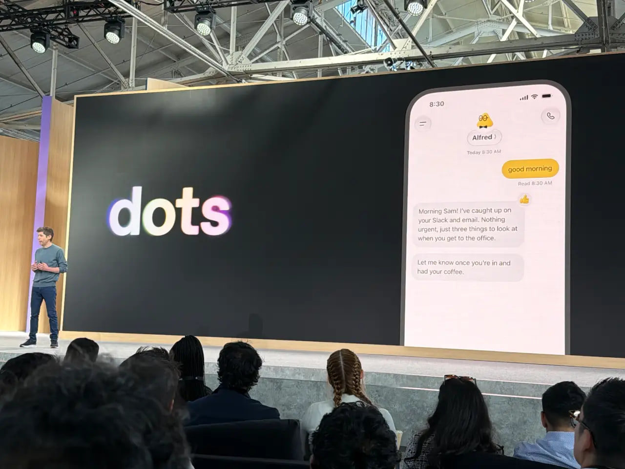 一位演讲者站在台上，观众席前，巨大的屏幕显示"dots"标志和一个模拟手机对话，对话中的助手名叫 Alfred。消息内容："早上好""早上好 Sam！我已经浏览了你的 Slack 和邮件。没有什么紧急的，只有三件事等你到办公室后看看""到了喝完咖啡后告诉我一声"