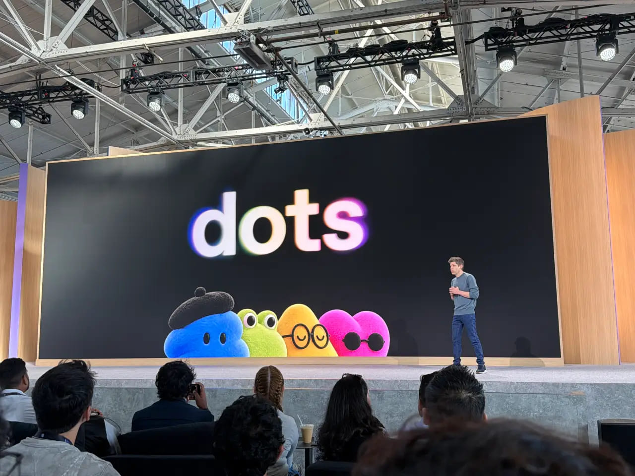 一位演讲者站在台上，观众席前，巨大的屏幕显示"dots"字样和四个彩色卡通角色。活动在一个大型室内场所，屋顶桁架外露，舞台灯光璀璨