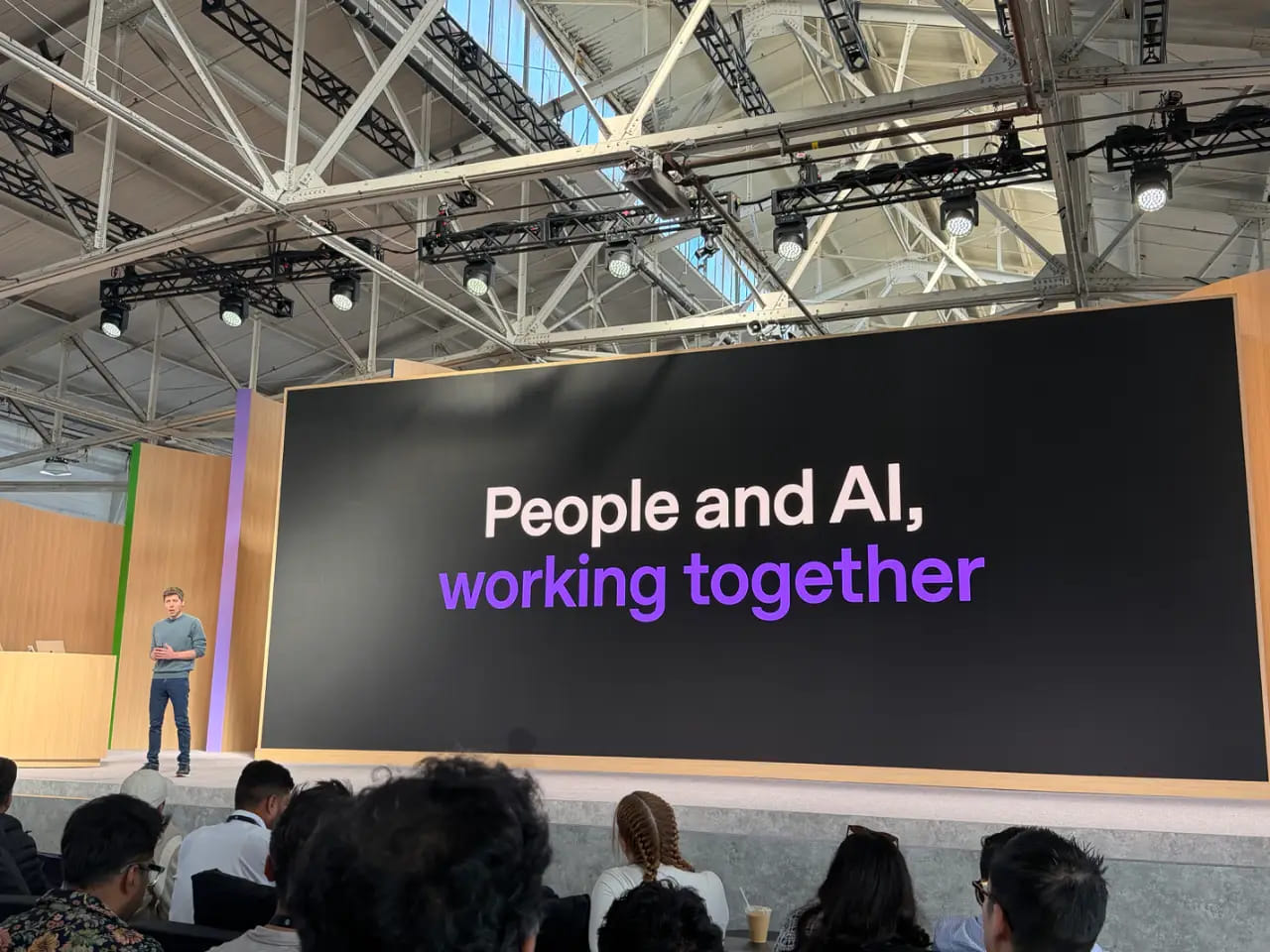 Sam Altman 站在会议舞台上，观众席前，巨大的屏幕显示"People and AI, working together"