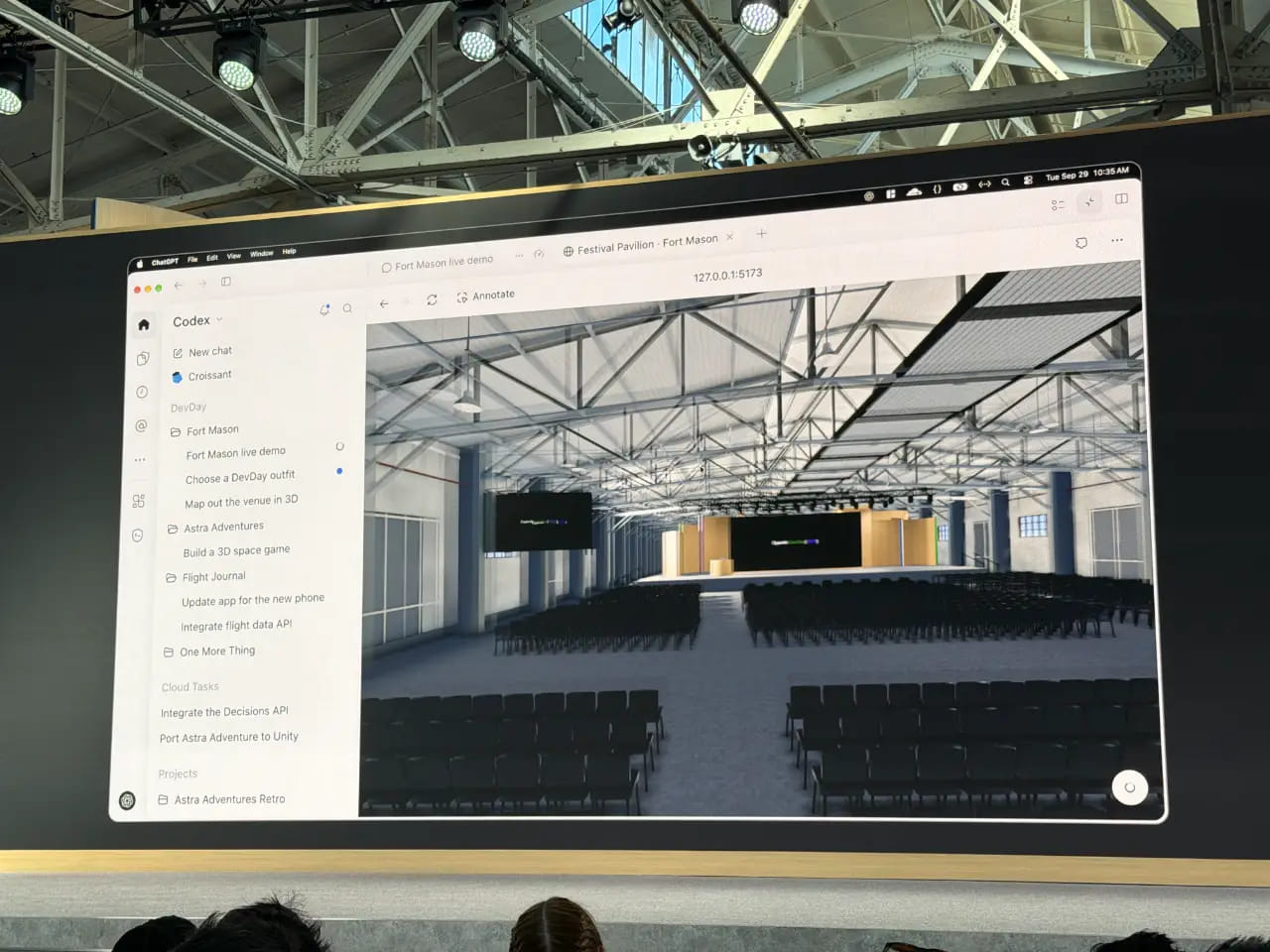 OpenAI DevDay 展区的一台大型显示器展示了一张计算机生成的 Fort Mason 节日馆视图，馆内布置了会议用的椅子排，面向舞台，屋顶桁架外露。Codex 侧边栏列出了项目，包括"Fort Mason 现场演示"和"用 3D 绘制场地地图"；底部可见观众们的头部